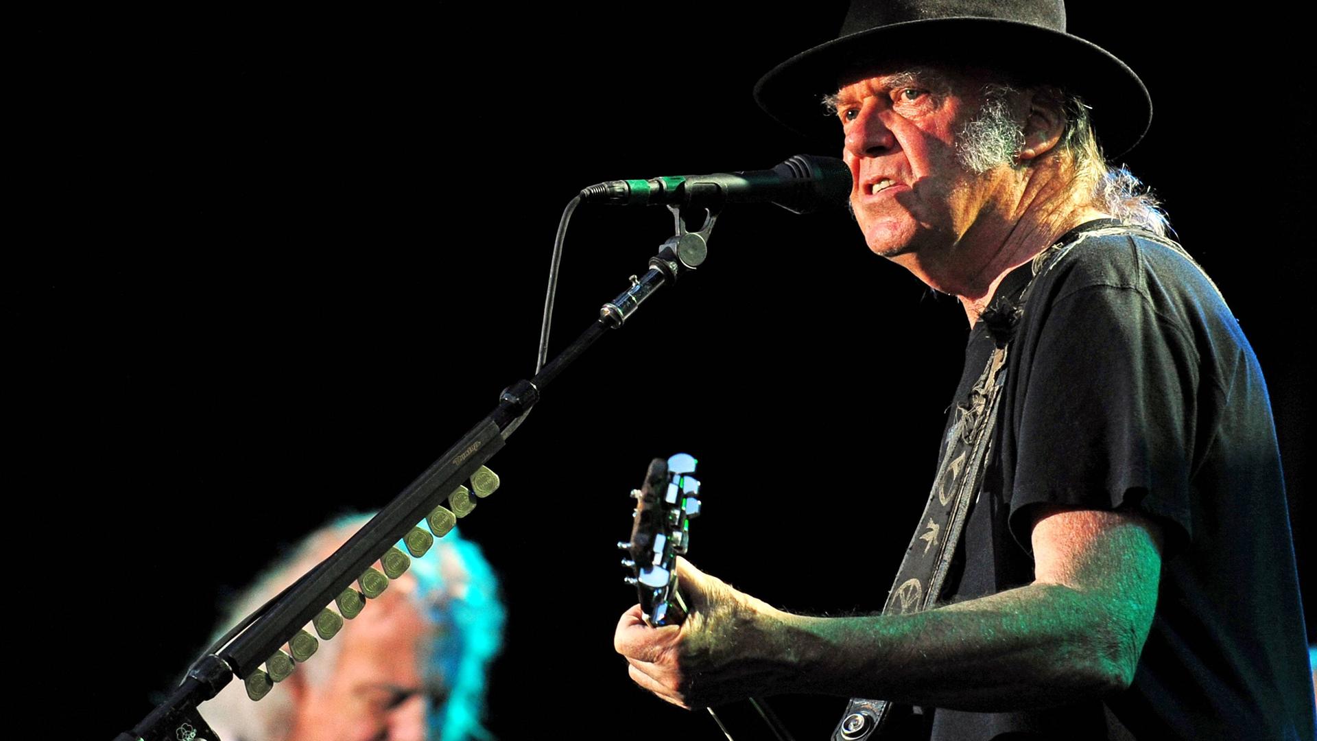 Neil Young auf einem Konzert mit seiner Band Crazy Horse im Jahr 2014. Young ist rechts auf dem Bild im Halbprofil zu sehen. Er hat einen schwarzen Hut auf, er spielt Gitarre. Neil Young auf einem Konzert mit seiner Band Crazy Horse im Jahr 2014. Young ist rechts auf dem Bild im Halbprofil zu sehen. Er hat einen schwarzen Hut auf, er spielt Gitarre.