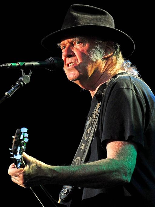 Neil Young auf einem Konzert mit seiner Band Crazy Horse im Jahr 2014. Young ist rechts auf dem Bild im Halbprofil zu sehen. Er hat einen schwarzen Hut auf, er spielt Gitarre.