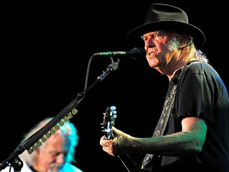Neil Young auf einem Konzert mit seiner Band Crazy Horse im Jahr 2014. Young ist rechts auf dem Bild im Halbprofil zu sehen. Er hat einen schwarzen Hut auf, er spielt Gitarre.