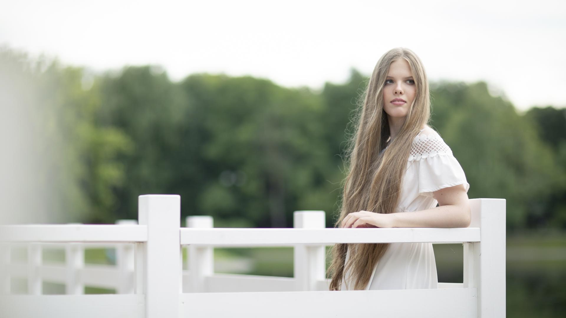 Eine junge Frau mit sehr langen blonden Haaren lehnt an eine weiße Brüstung. Sie trägt ein weißes Sommerkleid und blickt versonnen. Hinter ihr sind verschwommen Bäume zu erkennen. Eine junge Frau mit sehr langen blonden Haaren lehnt an eine weiße Brüstung. Sie trägt ein weißes Sommerkleid und blickt versonnen. Hinter ihr sind verschwommen Bäume zu erkennen.