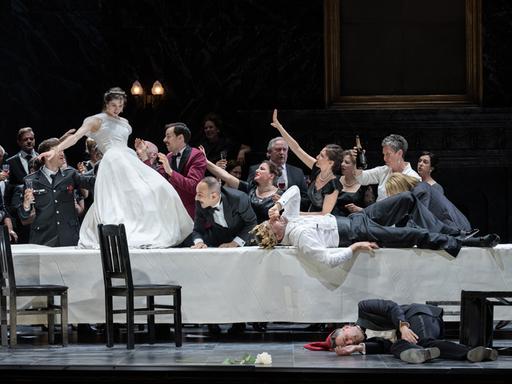 Auf einem Tisch steht eine Frau mit einem langen, weißen Kleid umgeben von Feiernden 