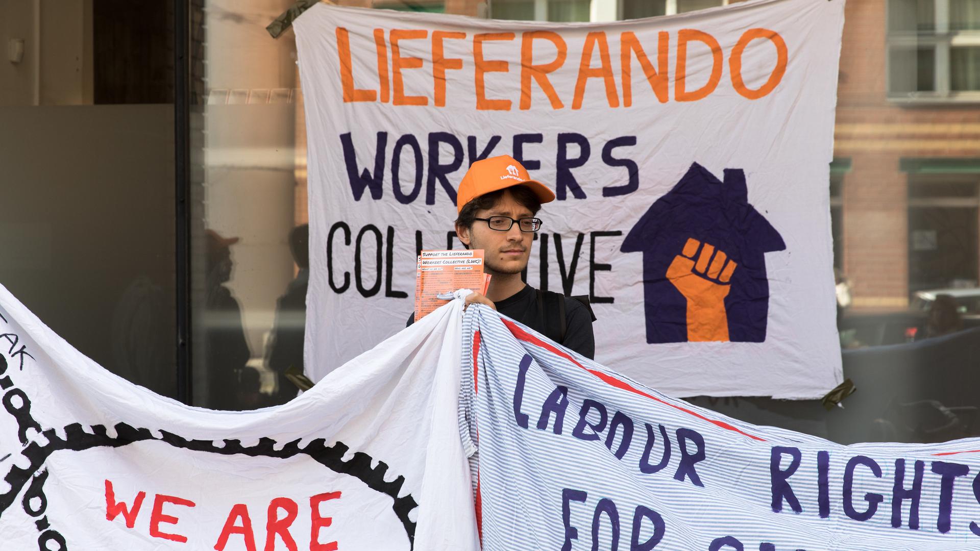 Ein Mitarbeiter des Lieferdienstes Lieferando inmitten von Protestplakaten bei einer Kundgebung in Berlin. Ein Mitarbeiter des Lieferdienstes Lieferando inmitten von Protestplakaten bei einer Kundgebung in Berlin.