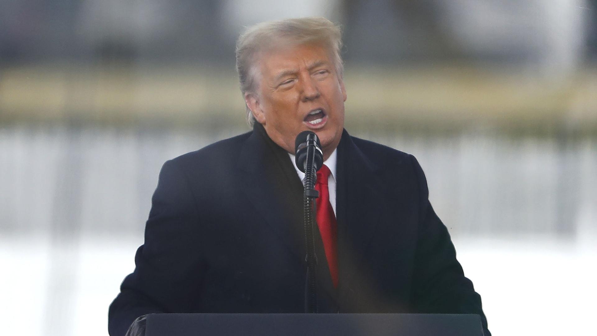 Trump steht im dunklen Wintermantel und mit roter Krawatte an einem Rednerpult mit dem Präsidentensiegel und sptricht in ein Mikrofon.