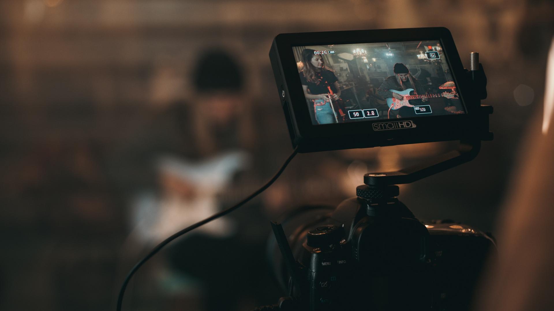 Zu sehen ist eine Kamera mit hochgeklapptem großem Display. Das Display zeigt zwei Musikerinnen mit Mikrofon und Bass, die vor der Kamera für ein Video spielen. Zu sehen ist eine Kamera mit hochgeklapptem großem Display. Das Display zeigt zwei Musikerinnen mit Mikrofon und Bass, die vor der Kamera für ein Video spielen.