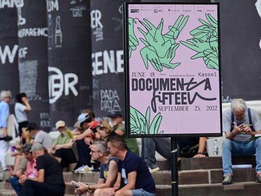 Besucher der documenta fifteen sitzen auf den Stufen des Fridericianums vor bemalten Säulen des Gebäudes; rechts ein Plakat der documenta