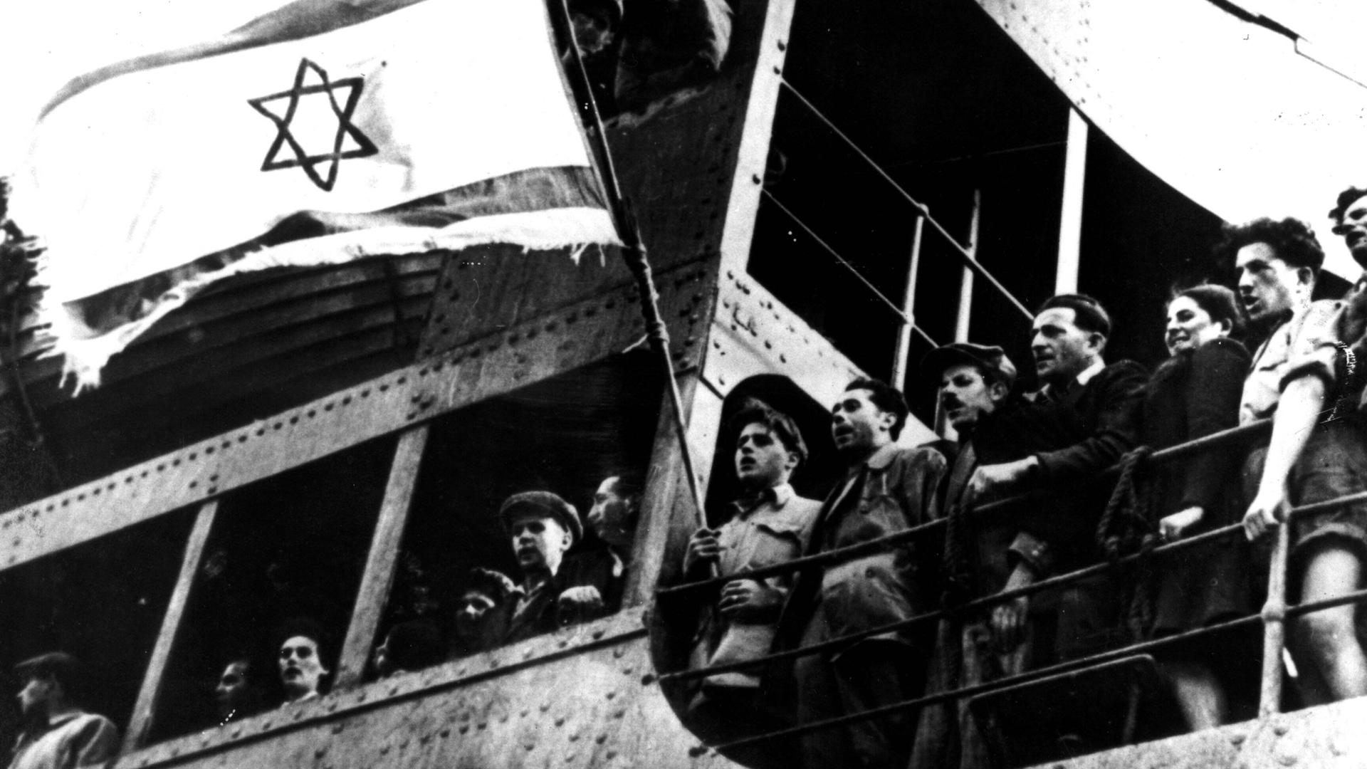 Schwarzweißaufnahme aus Untersicht von jüdischen Flüchtlingen, die von einem Schiff aus an Land blicken und dabei eine Flagge mit Davidstern schwenken.