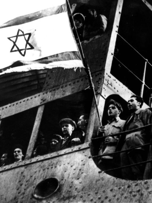Schwarzweißaufnahme aus Untersicht von jüdischen Flüchtlingen, die von einem Schiff aus an Land blicken und dabei eine Flagge mit Davidstern schwenken.