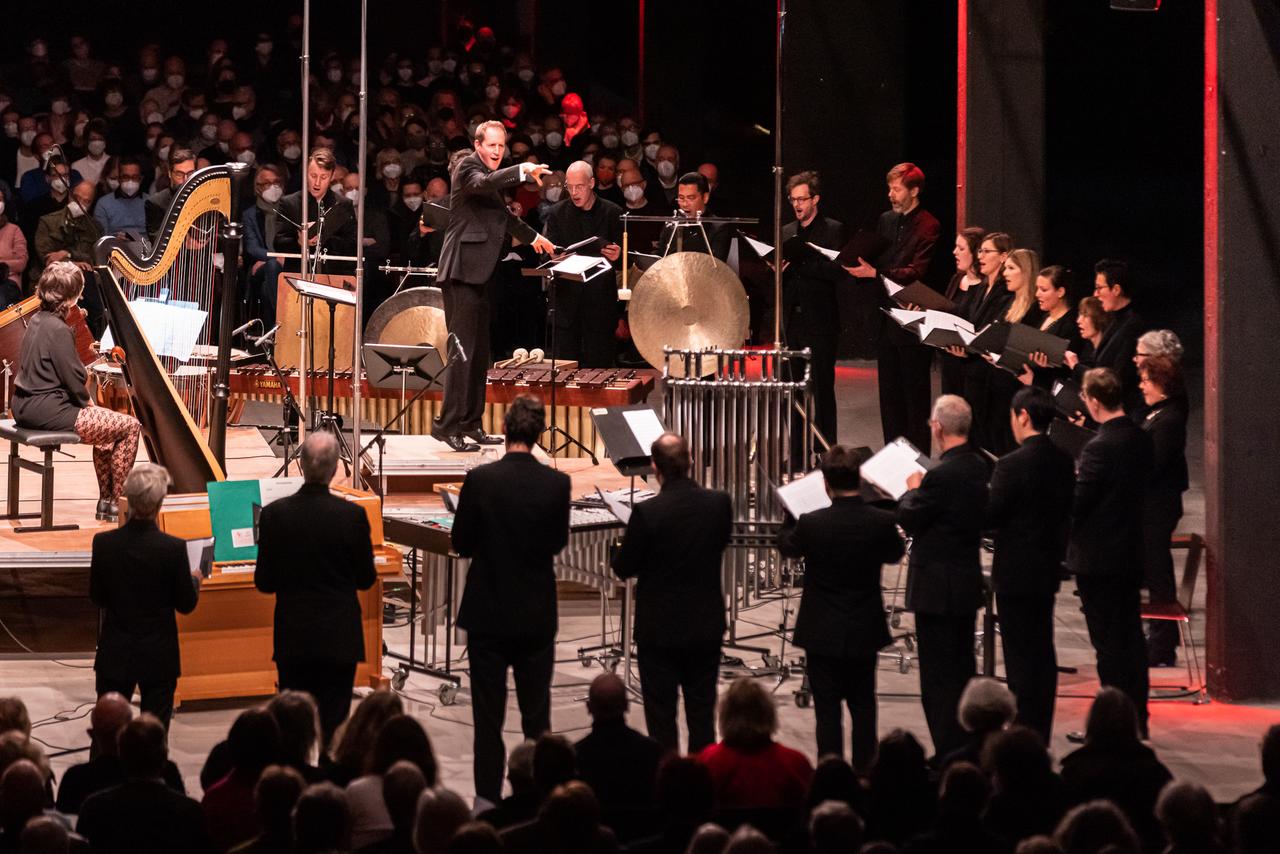 Der Chor steht um verschiedene Schlagwerke und andere Instrumente gruppiert und singt in Richtung des Chefdirigenten Justin Doyle. Der Chor steht um verschiedene Schlagwerke und andere Instrumente gruppiert und singt in Richtung des Chefdirigenten Justin Doyle.