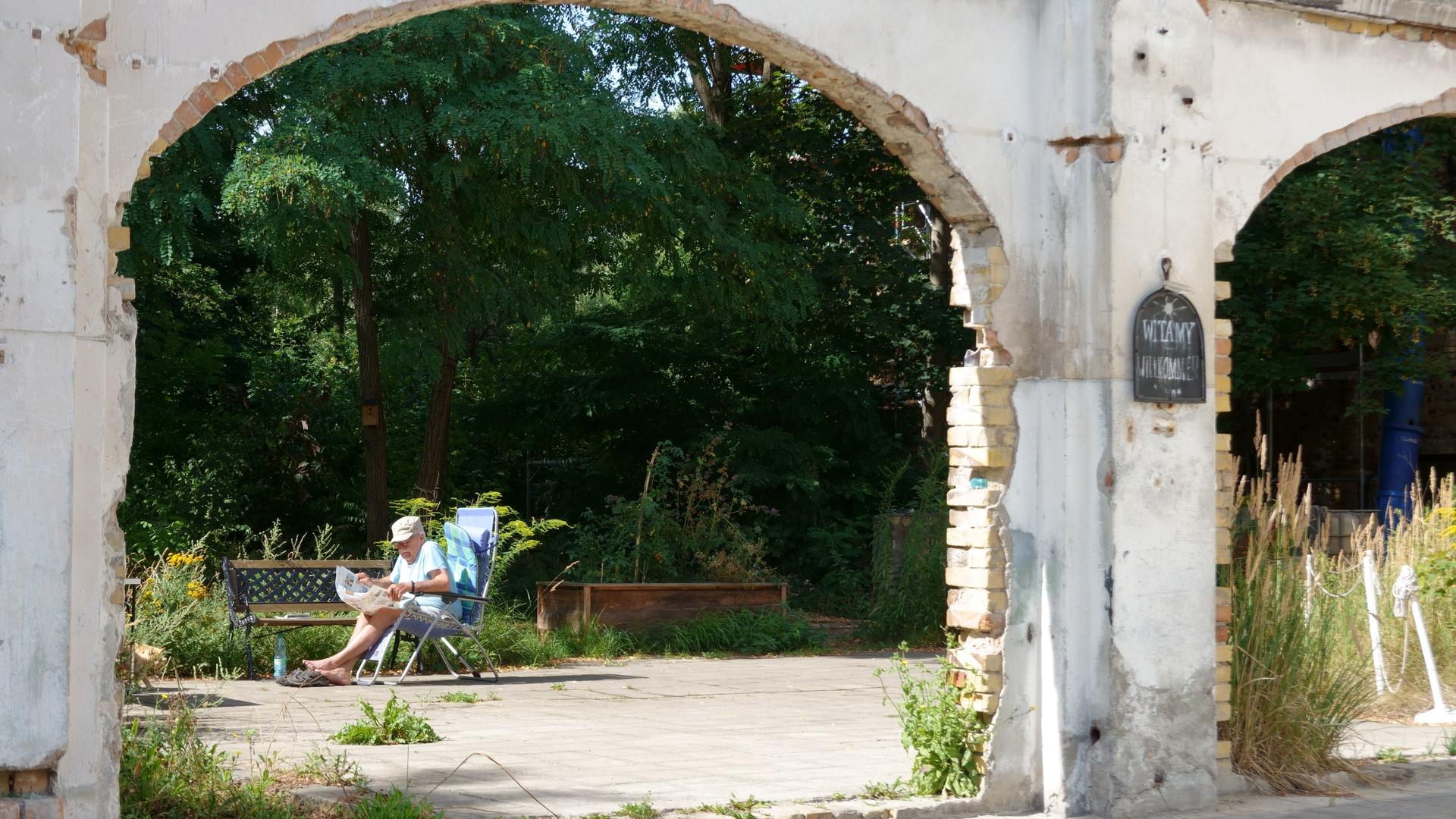 Ein Rentner sitzt auf einem Gartenstuhl und liest Zeitung. Man sieht ihn durch einen ausgefranzten Torbogen durch.