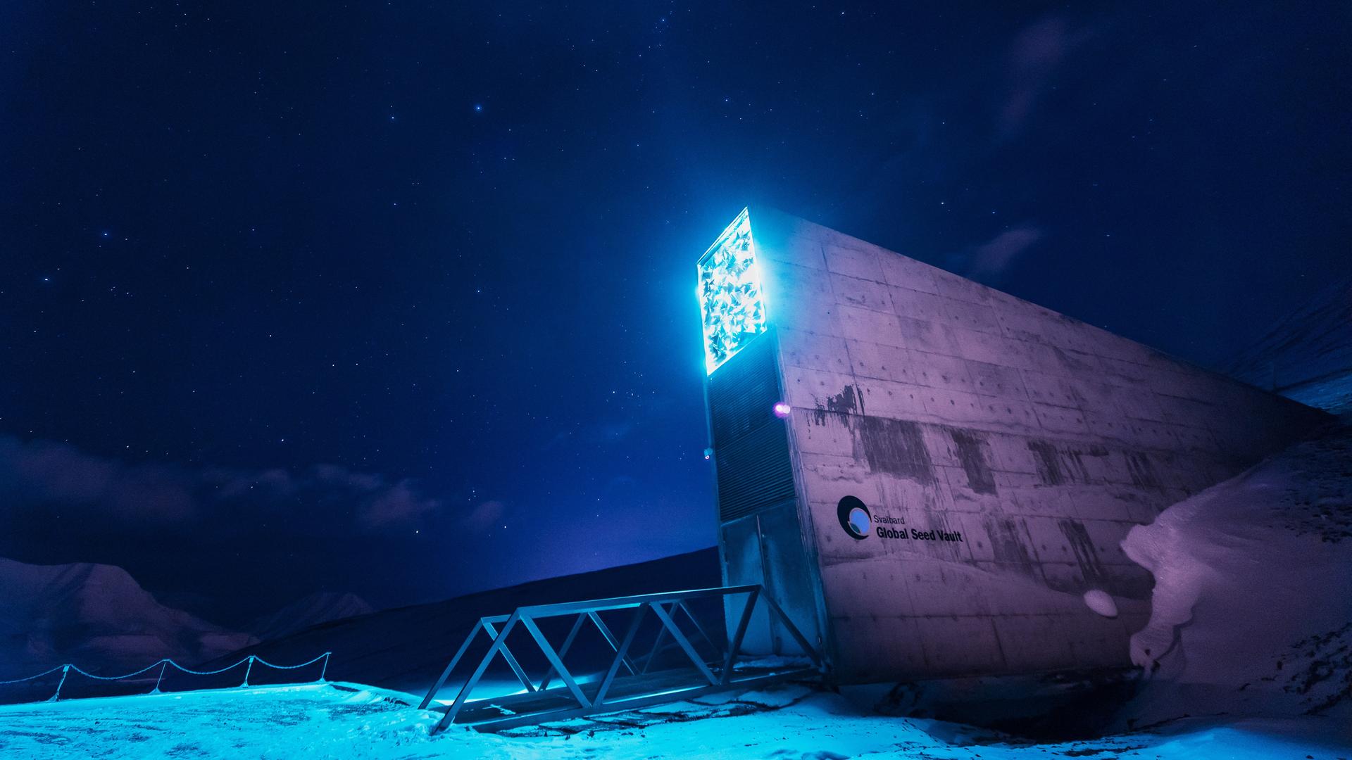 Aufragender Eingang zu einem unteridisch liegenden Gebäude mit der Aufschrift "Global Seed Vault". Seine Beleuchtung hebt es vor dem Sternenhimmel ab. Aufragender Eingang zu einem unteridisch liegenden Gebäude mit der Aufschrift "Global Seed Vault". Seine Beleuchtung hebt es vor dem Sternenhimmel ab.