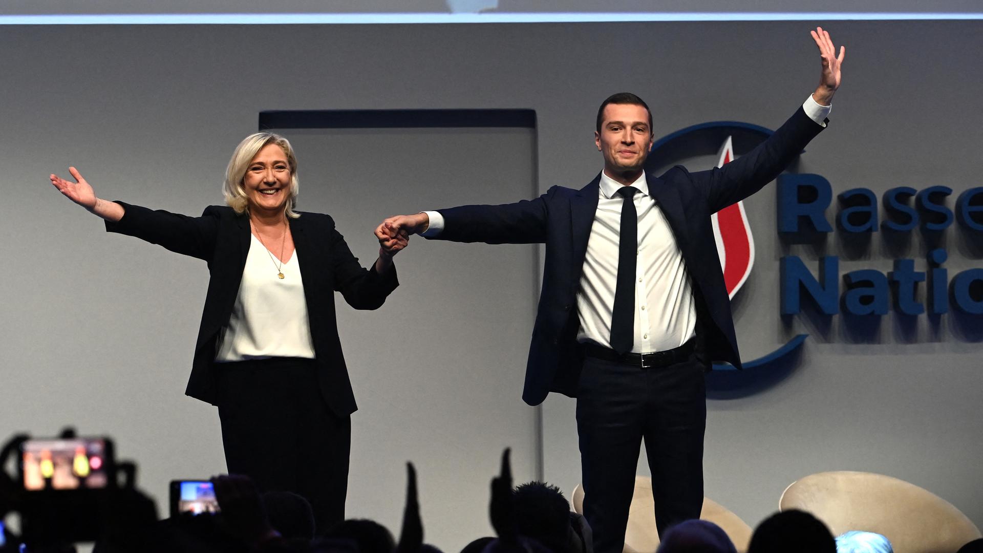 Die Fraktionsvorsitzende der französischen rechtsradikalen Partei Rassemblement National (RN), Marine Le Pen, und der neu gewählte Parteivorsitzende Jordan Bardella stehen auf einer Bühne, halten sich an der Hand und winken ins Publikum.