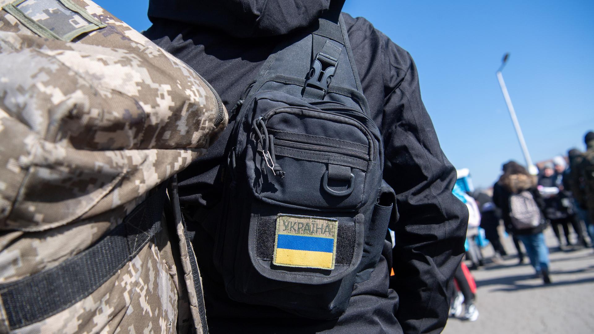 Menschen am polnischen Grenzübergang zur Ukraine, im Vordergrund ein Rücken mit Tarnfleck-Rucksack und einer Tasche mit Ukraine-Flaggenaufnäher Menschen am polnischen Grenzübergang zur Ukraine, im Vordergrund ein Rücken mit Tarnfleck-Rucksack und einer Tasche mit Ukraine-Flaggenaufnäher