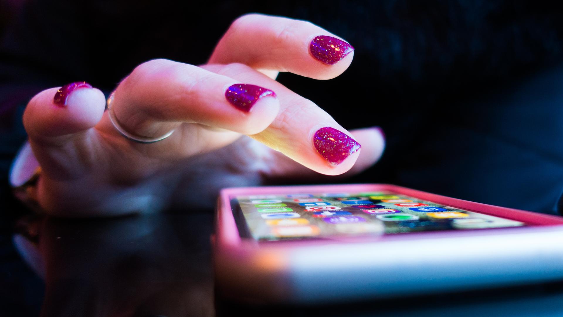 Nahaufnahme einer weiblichen Hand mit glitzernd roten Fingernägeln schwebt zögerlich über dem Bildschirm eines Smartphones. Nahaufnahme einer weiblichen Hand mit glitzernd roten Fingernägeln schwebt zögerlich über dem Bildschirm eines Smartphones.