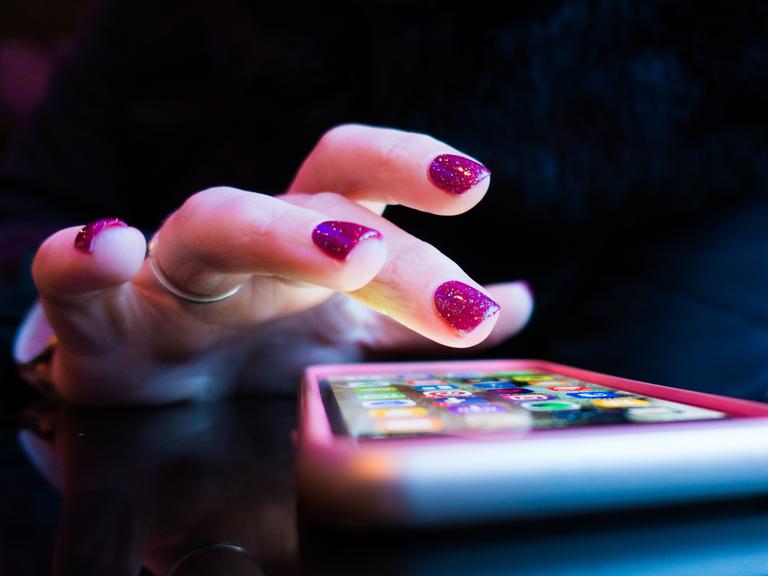 Nahaufnahme einer weiblichen Hand mit glitzernd roten Fingernägeln schwebt zögerlich über dem Bildschirm eines Smartphones.