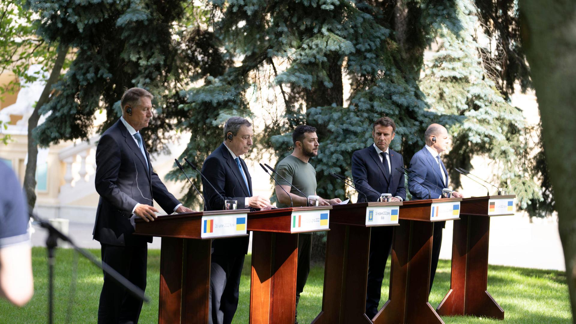 Fünf Männer stehen in einem Garten vor Rednerpulten und geben eine Pressekonferenz. 