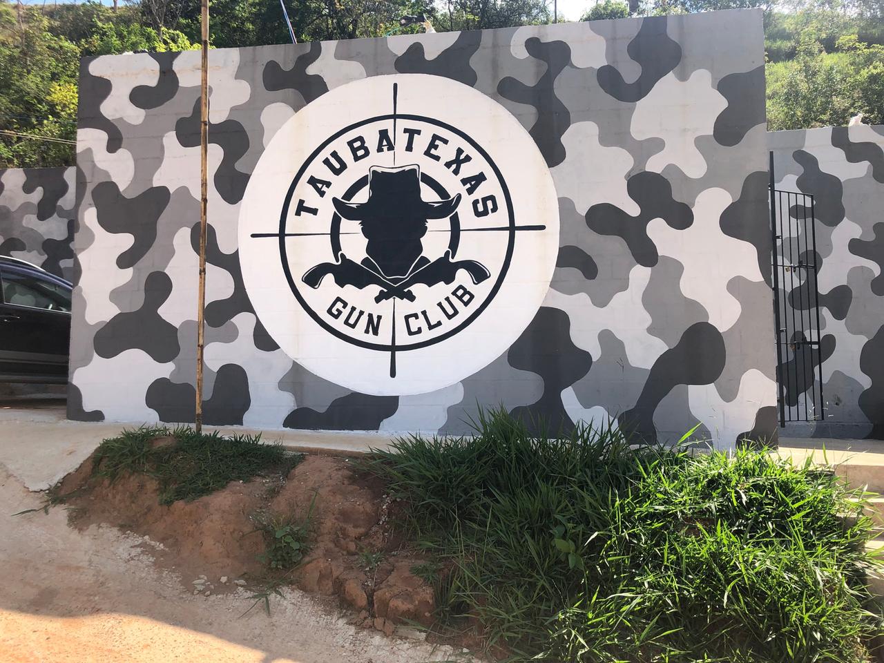 An einer Wand in grauen Tarnfarben prangt das Logo des Taubatexas Gun Club: Die schwarze Silhouette eines Mannes mit Hut im Fadenkreuz. An einer Wand in grauen Tarnfarben prangt das Logo des Taubatexas Gun Club: Die schwarze Silhouette eines Mannes mit Hut im Fadenkreuz.