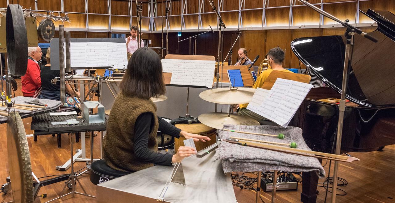 Blick hinter der Schlagwerkerin, die eine Saite auf einem tischartigen Instrument streicht, zum Dirigenten, den weitere Musiker beim Spiel umgeben. Blick hinter der Schlagwerkerin, die eine Saite auf einem tischartigen Instrument streicht, zum Dirigenten, den weitere Musiker beim Spiel umgeben.