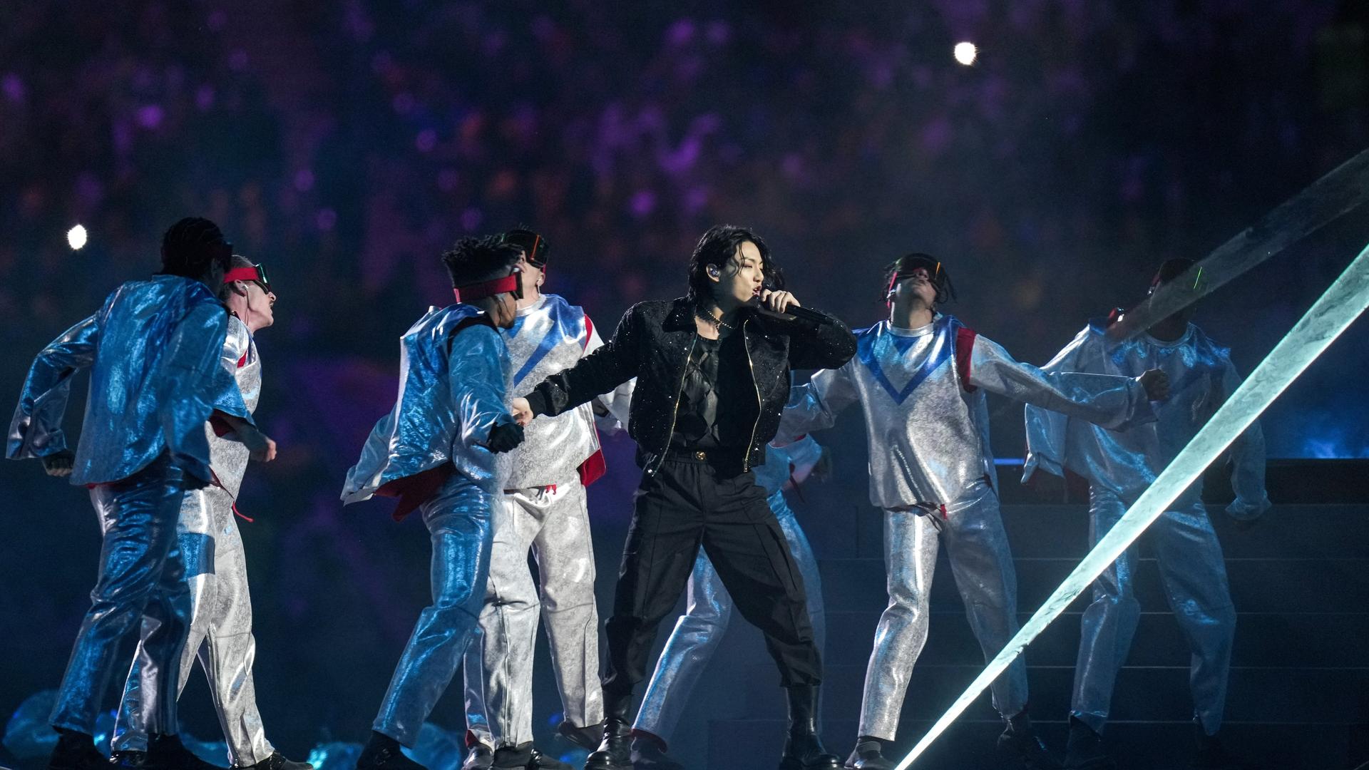 Der koreanische Sänger und Popstar Jungkook steht auf einer Bühne im WM-Stadion in Katar und singt. Hinter ihm sind die Tänzer der Bühnenshow zu sehen. 