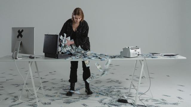 Eine Frau steht an einem Tisch und holt Geldscheine aus einem Koffer und scheint sie zählen zu wollen. Auf bem Boden sind auch Geldscheine verteilt.