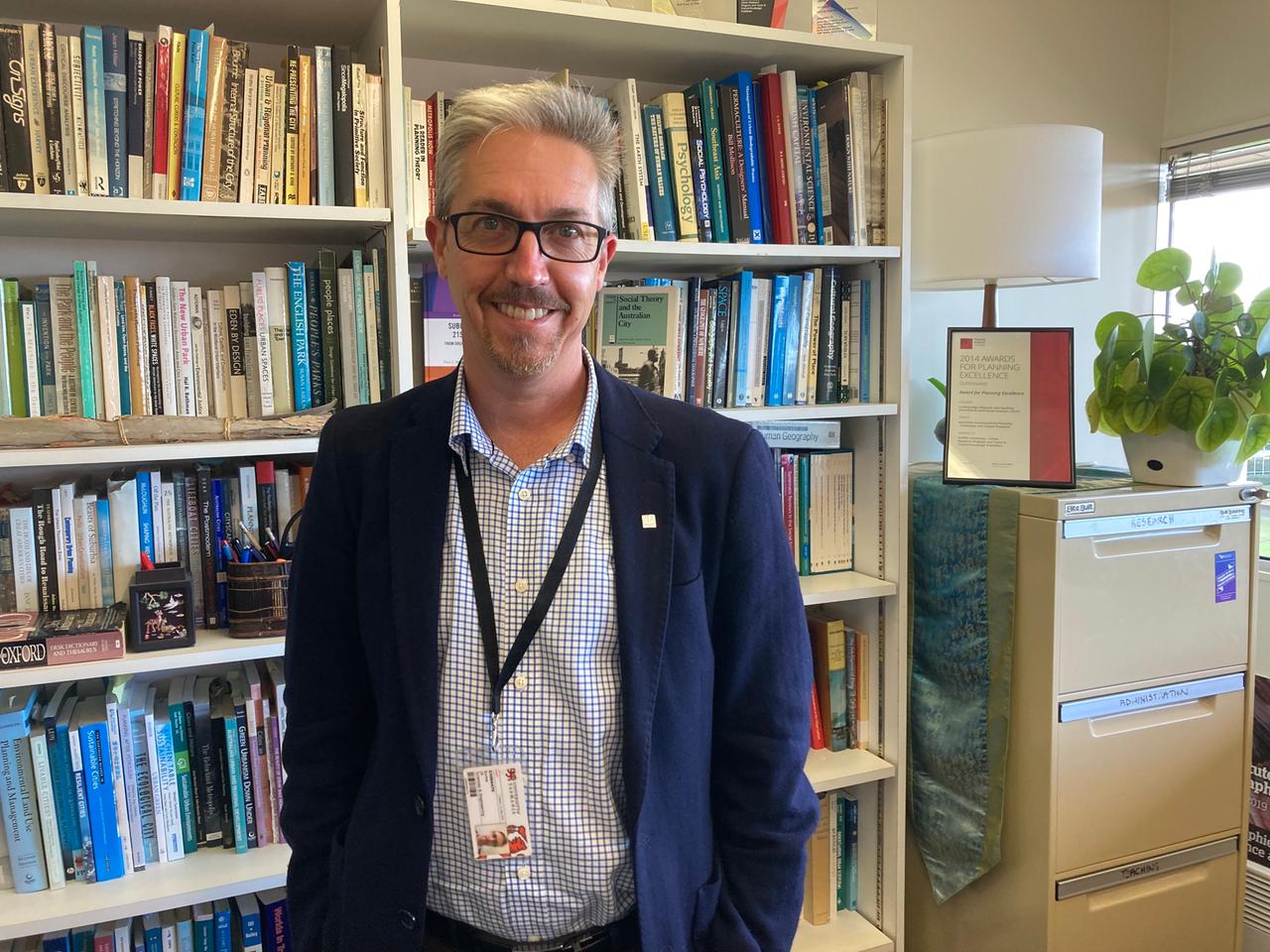 Ein Mann im blauen Jacket und kariertem Hemd, mit Brille und kurzem grauen Haar steht vor einer Bücherwand. Ein Mann im blauen Jacket und kariertem Hemd, mit Brille und kurzem grauen Haar steht vor einer Bücherwand.