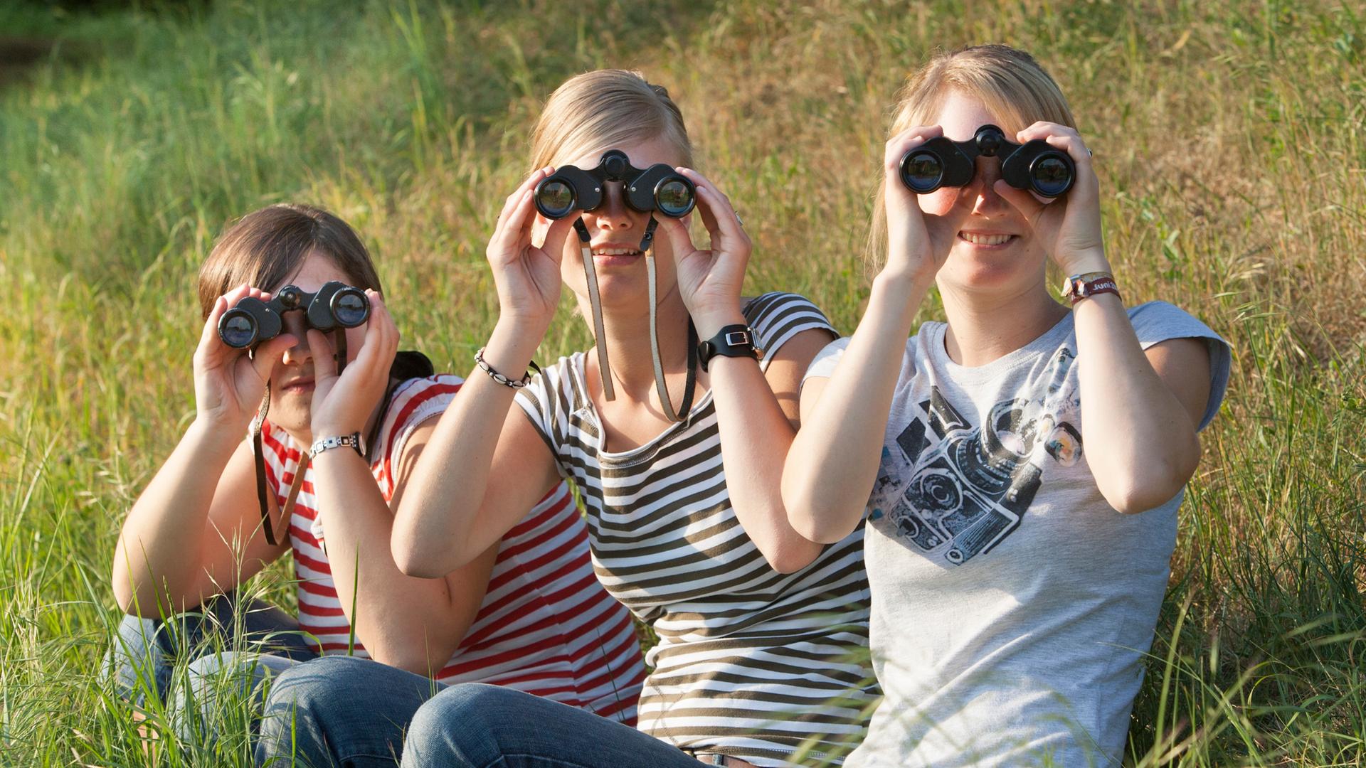 Junge Frauen sitzen auf einer Wiese und schauen durch Fern-Gläser.