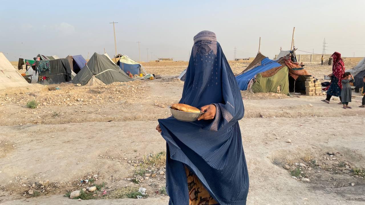 Eine Frau trägt eine blaue Burka, die auch ihr Gesicht verhüllt, in der Hand hält sie eine Schüssel mit Brot. Im Hintergrund: Zelte in einer staubigen Landschaft.