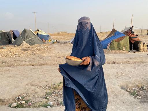 Eine Frau trägt eine blaue Burka, die auch ihr Gesicht verhüllt, in der Hand hält sie eine Schüssel mit Brot. Im Hintergrund: Zelte in einer staubigen Landschaft.