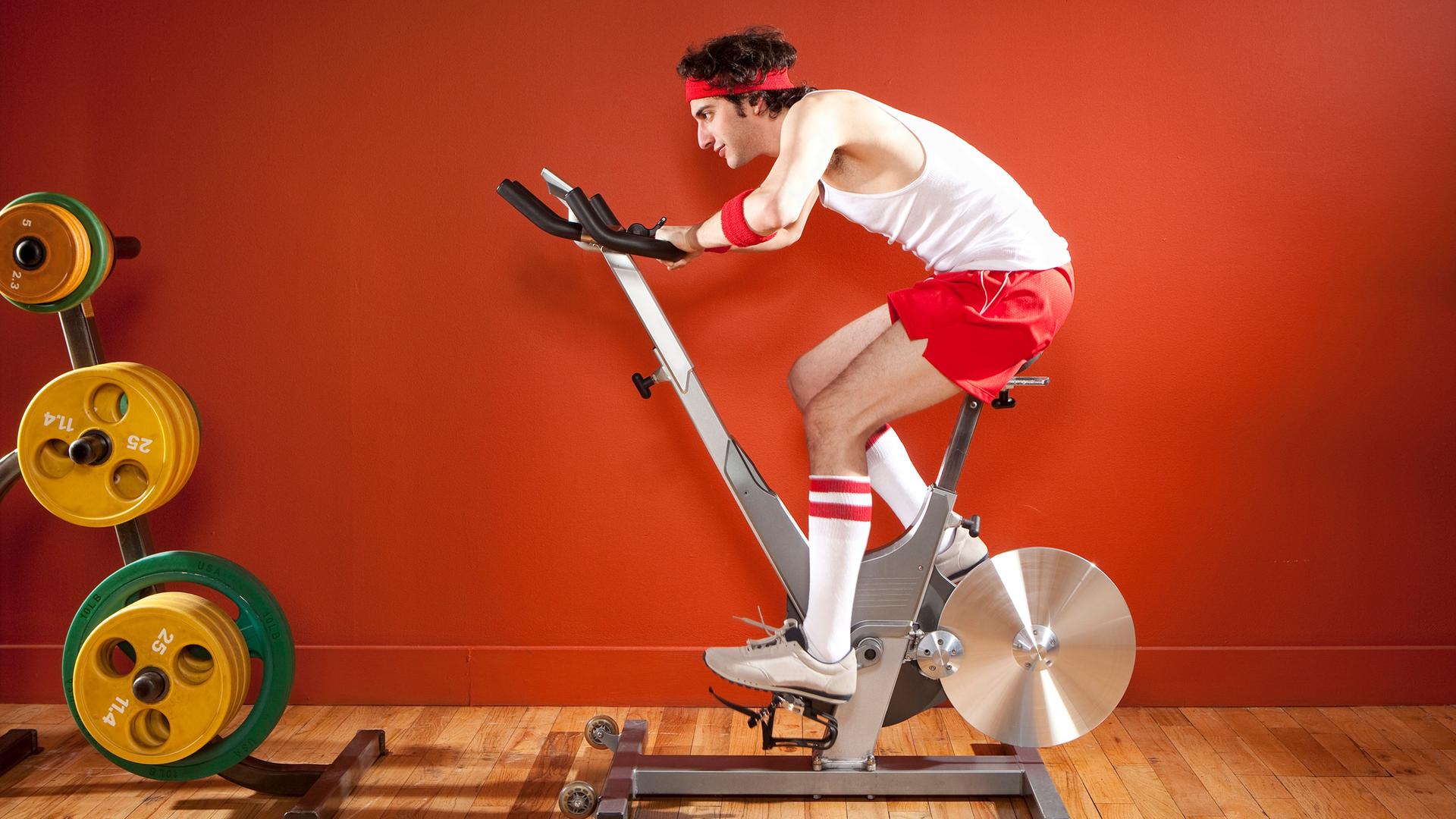 Ein Mann in einem Sportoutfit radelt auf einem Hometrainer. Die Wand hinter ihm ist rot. Vor ihm liegen Hanteln auf dem Boden. Ein Mann in einem Sportoutfit radelt auf einem Hometrainer. Die Wand hinter ihm ist rot. Vor ihm liegen Hanteln auf dem Boden.