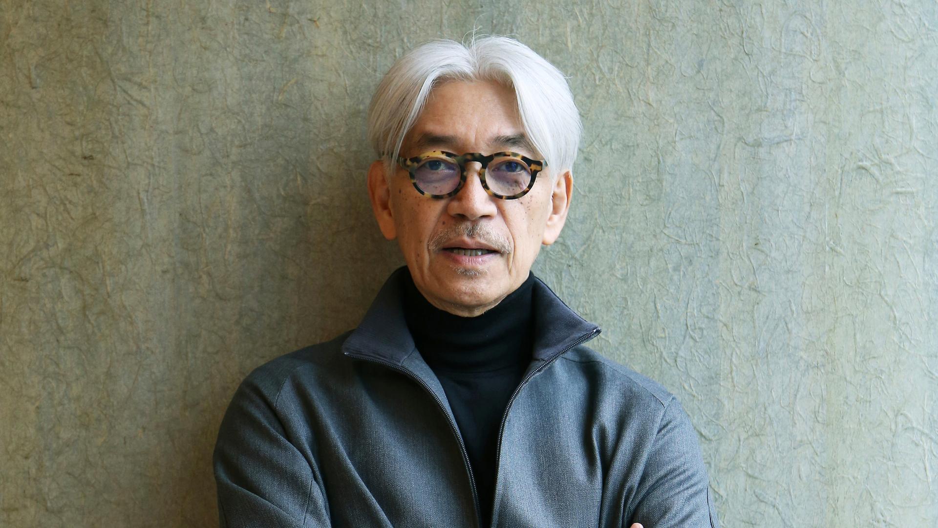Der japanische Komponist und Pianist Ryūichi Sakamoto mit auffällig dunkel gerahmter Brille und verschränkten Armen lehnt an einer grauen Wand.