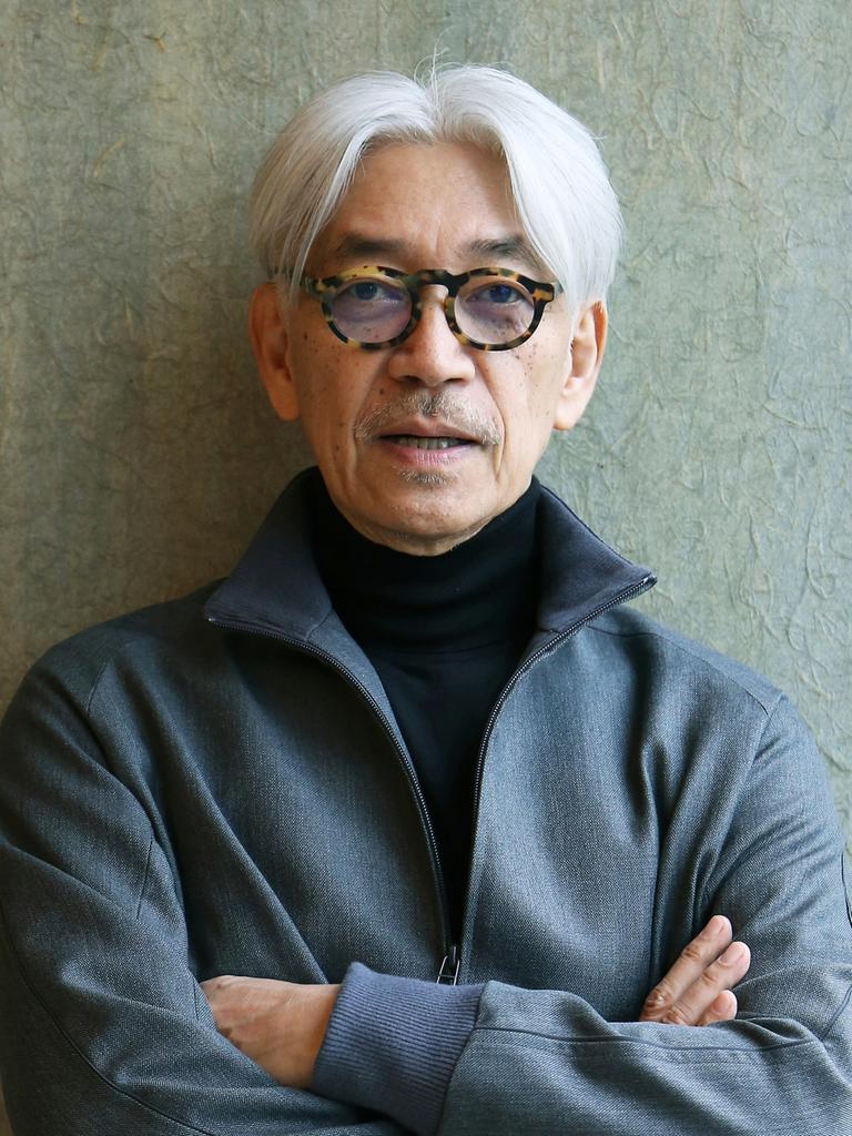Der japanische Komponist und Pianist Ryūichi Sakamoto mit auffällig dunkel gerahmter Brille und verschränkten Armen lehnt an einer grauen Wand.