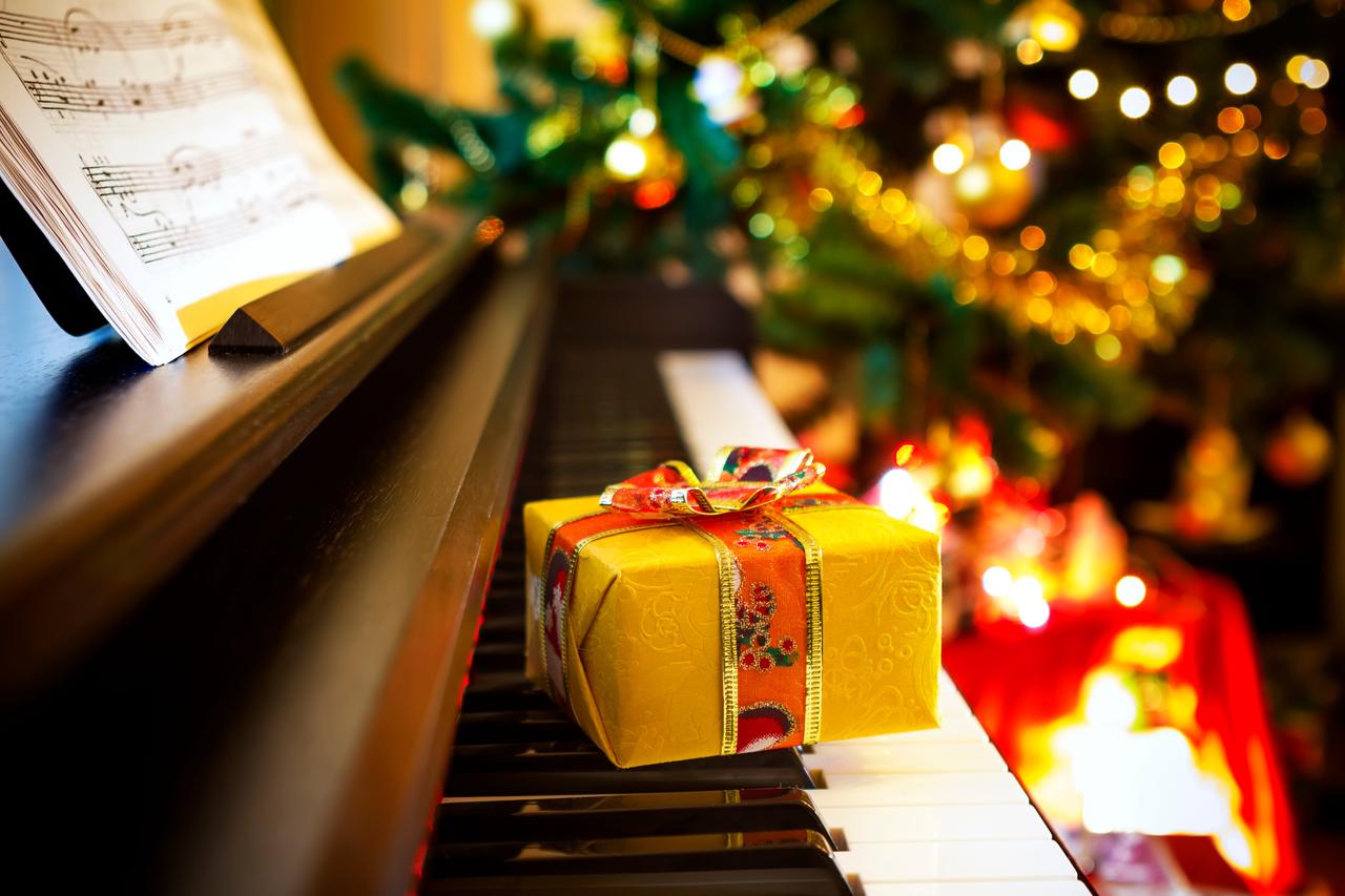 Ein kleines, gelbes Geschenkpäckchen mit großer Schleife liegt auf einer Klaviertastatur. Dahinter ist schemenhaft ein Weihnachtsbaum zu erkennen.