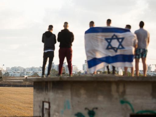 Mehrere Männer stehen auf einem Dach und haben eine große Israelflagge um sich geschlungen. Sie schauen in die Ferne.