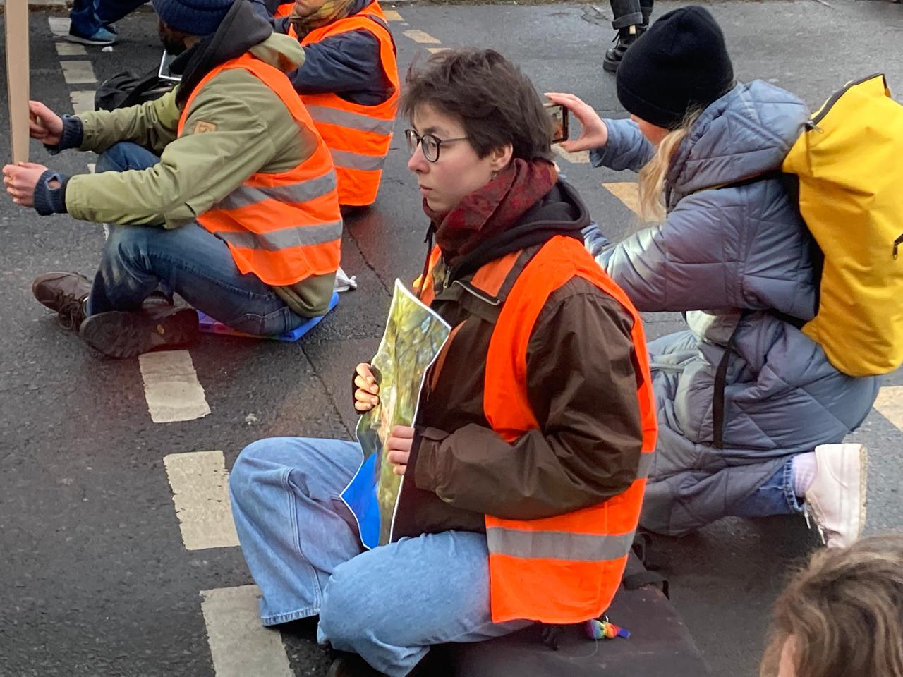 Die Aktivistin Lina Eichler von Die Letzte Generation sitzt in orangener Sicherheitsweste auf der Straße und hält ein Bild in der Hand.
