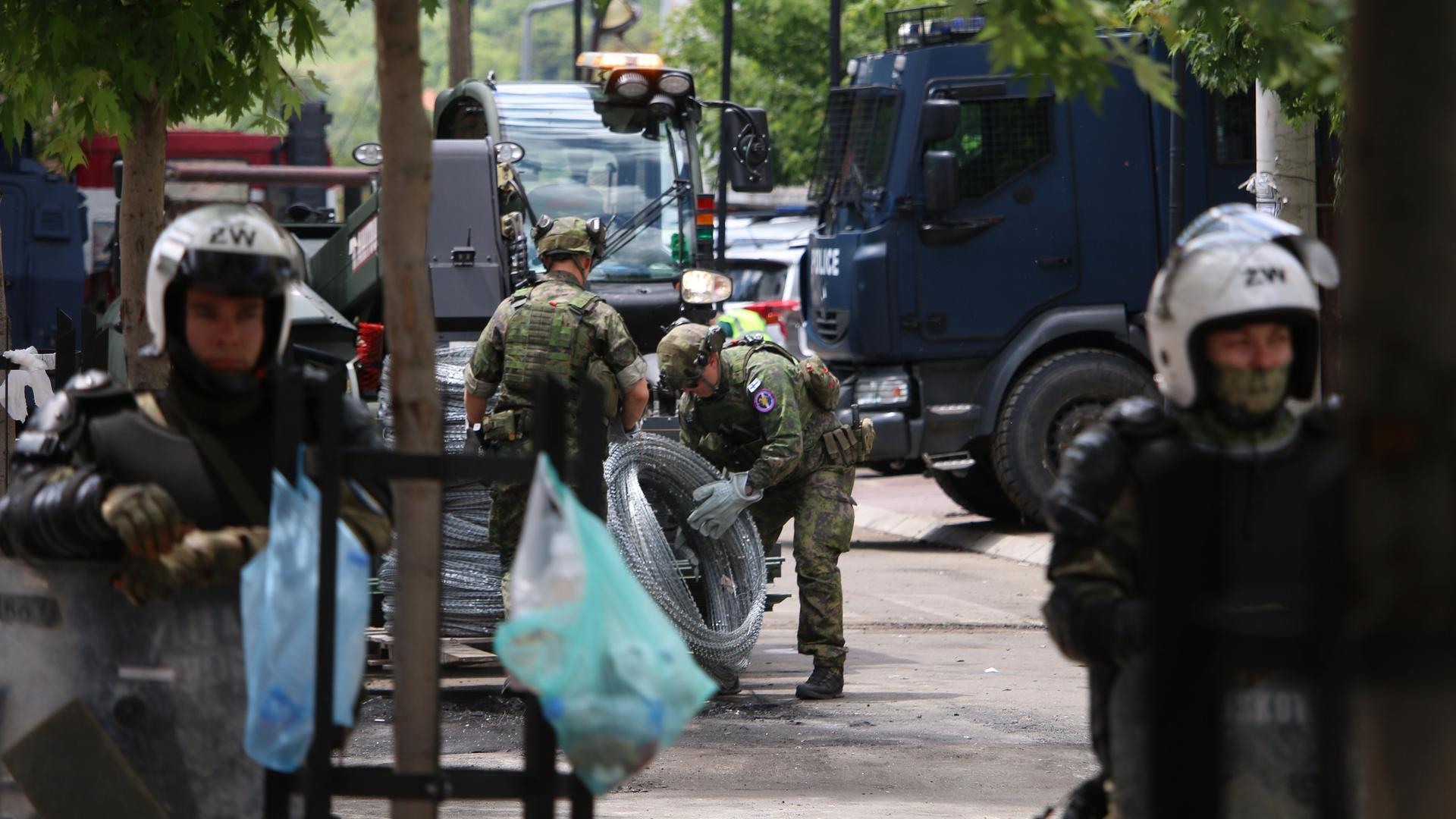 Zwischen  lokalen Polizeikräften sind KFOR Soldaten in Tarnuniformen zu sehen, die Natodraht ausrollen.