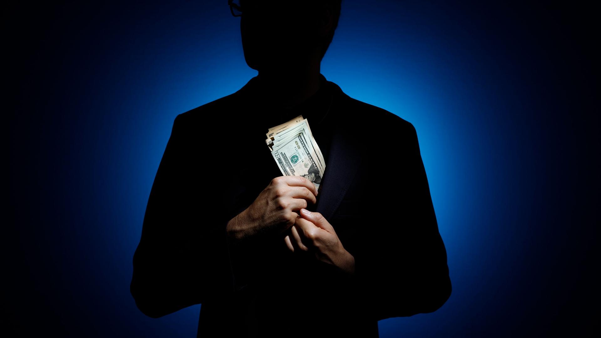 Silhouette eines Mannes vor blauem Licht, der sich ein Bündel Geldscheine in das Sakko steckt. Silhouette eines Mannes vor blauem Licht, der sich ein Bündel Geldscheine in das Sakko steckt.