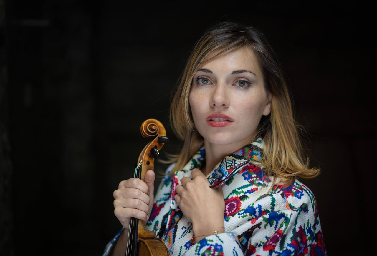 Diana Tishchenko hält in der einen Hand ihre Geige, in der anderen schlägt sie ihren Mantel zu, der ein Stickmuster zeigt. Diana Tishchenko hält in der einen Hand ihre Geige, in der anderen schlägt sie ihren Mantel zu, der ein Stickmuster zeigt.