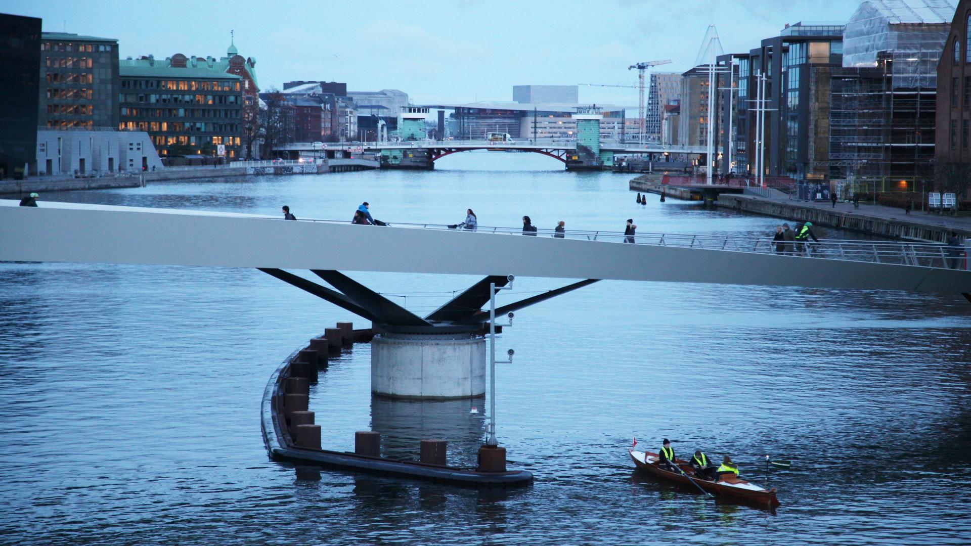 Radfahrer fahren über die Lille Langebro im Zentrum von Kopenhagen, während ein Ruderboot unter der Brücke hindurchfährt. 
