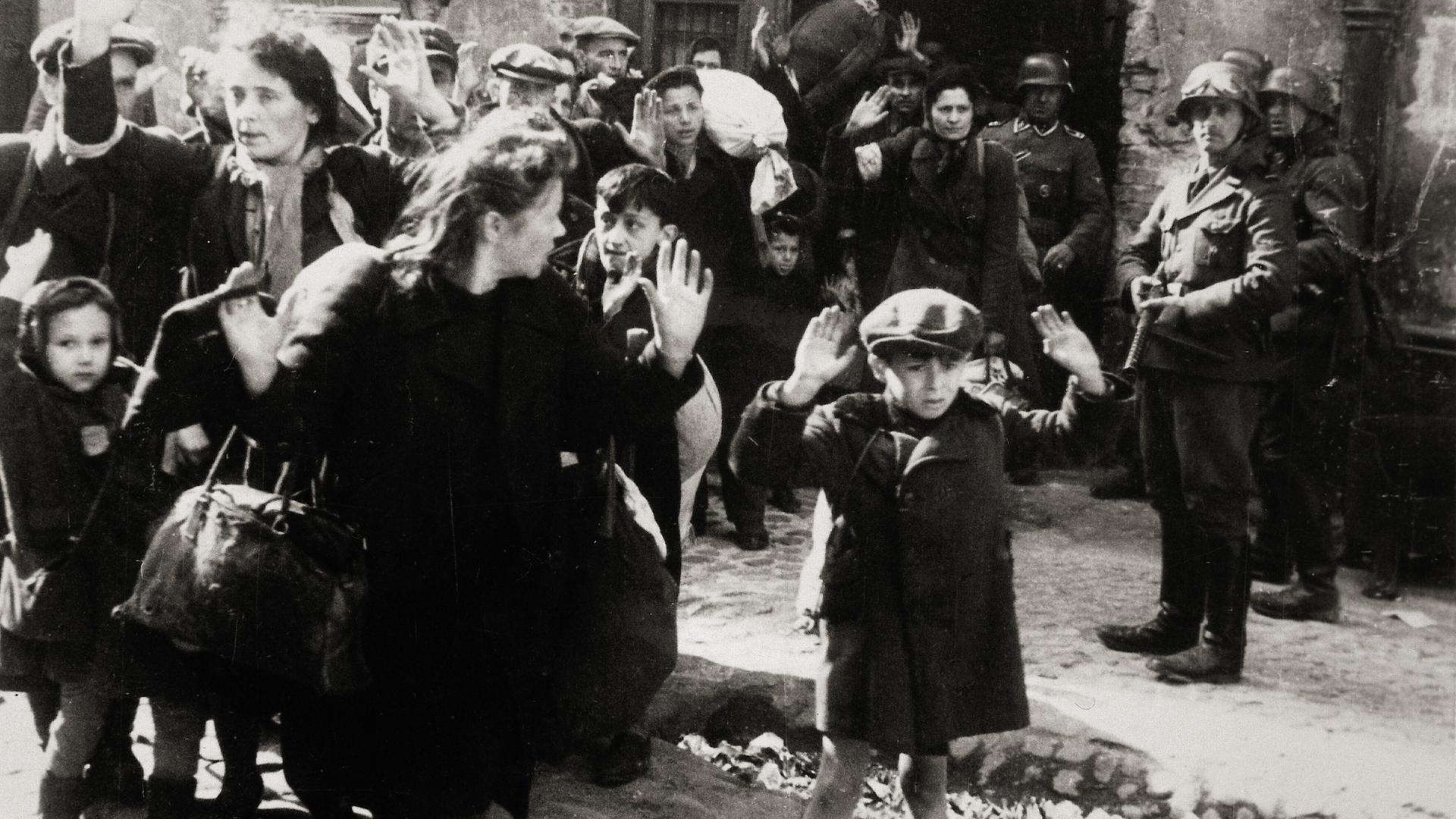 Warschauer Ghettoaufstand April/Mai 1943: Kleiner Junge mit erhobenen Händen, Frauen und andere Kinder kommen aus einem von den deutschen Truppen eroberten Haus.