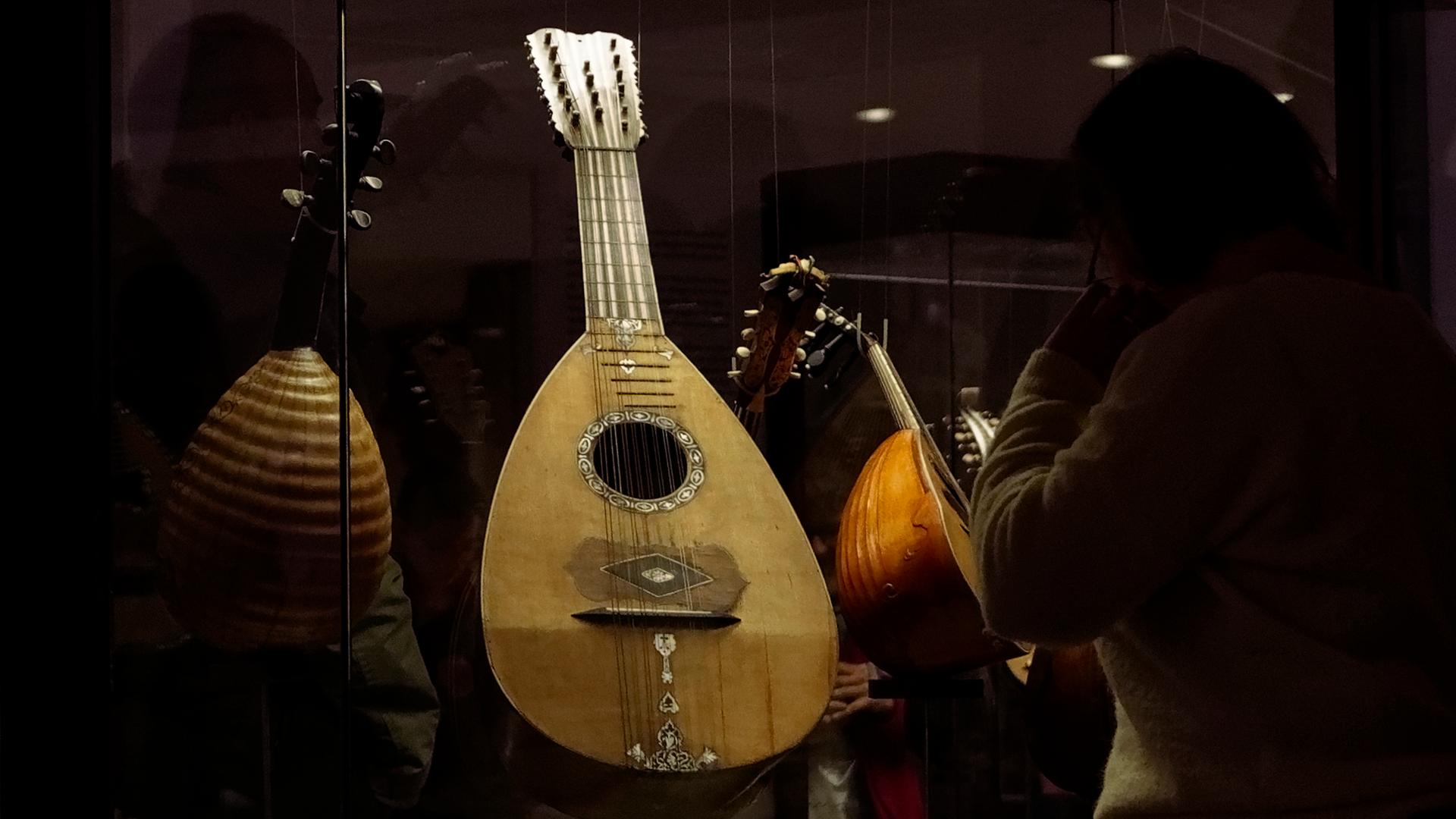 Ein Zupfinstrument aus hellem Holz steht in einer Vitrine. Der Hals des Instruments ist eher kurz, der Resonanzkörper rund, es ist eine Mandoline.