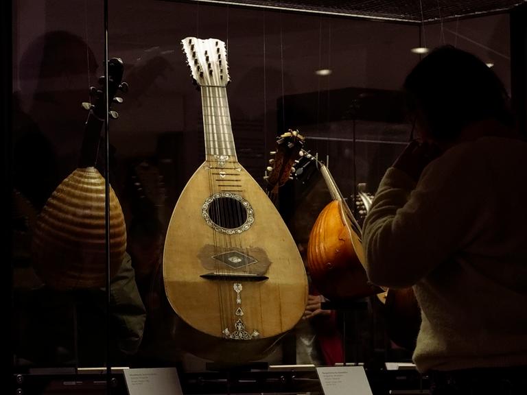 Ein Zupfinstrument aus hellem Holz steht in einer Vitrine. Der Hals des Instruments ist eher kurz, der Resonanzkörper rund, es ist eine Mandoline.