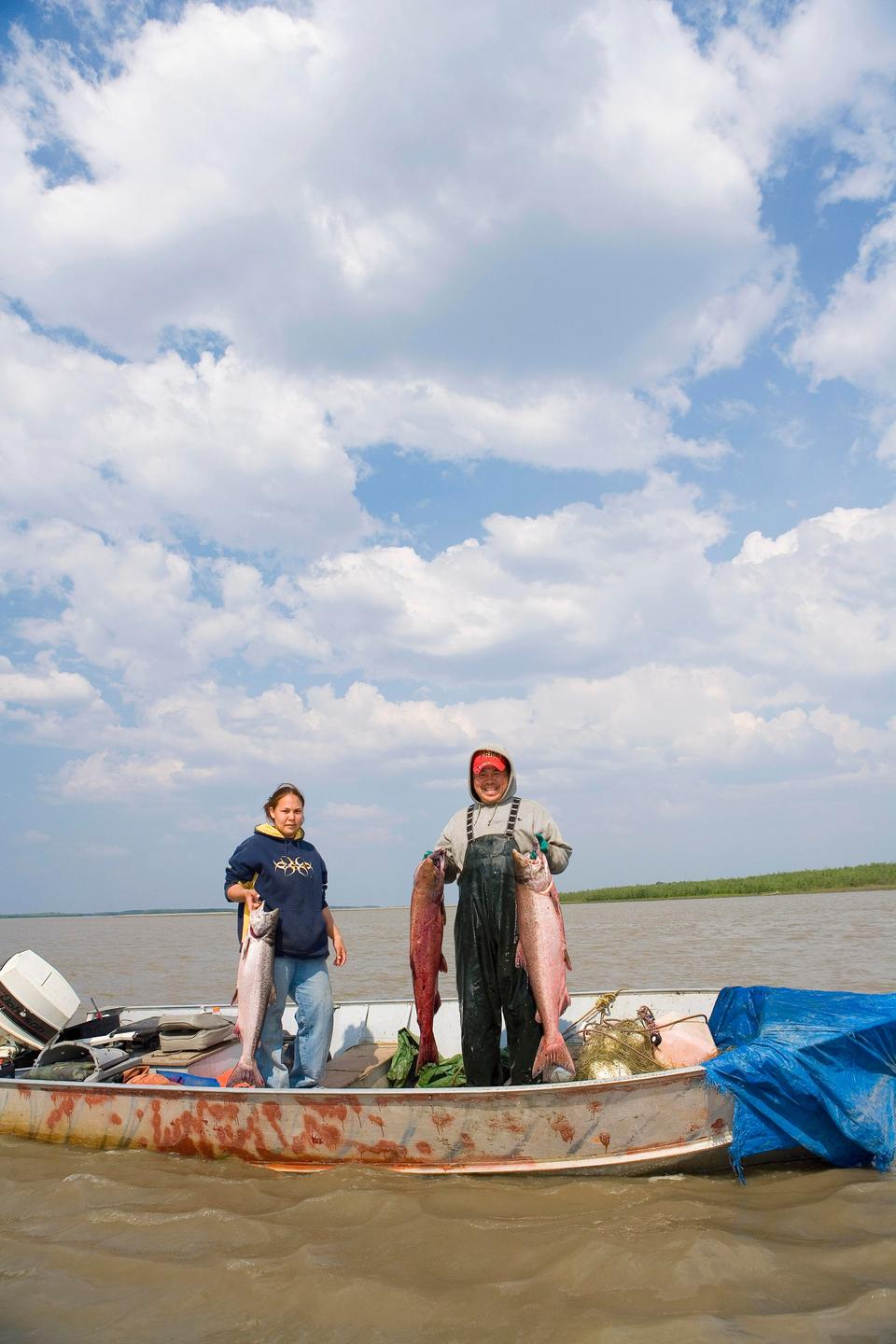 Zwei Personen stehen in Alaska in einem Boot und halten gefangene Fische hoch. 