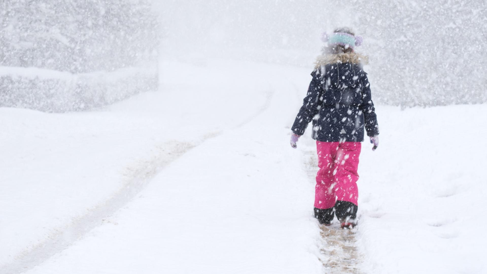 Ein Kind in einer pinken Schneehose, Handschuhen, Stirnband und dicker Jacke läuft durch Schneegestöber und hinterlässt dabei eine Spur in der Landschaft.