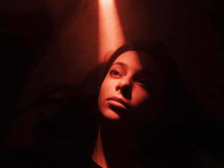 Symbolbild: Eine junge Frau in einem dunklen Raum mit einem Lichtschein von oben. 