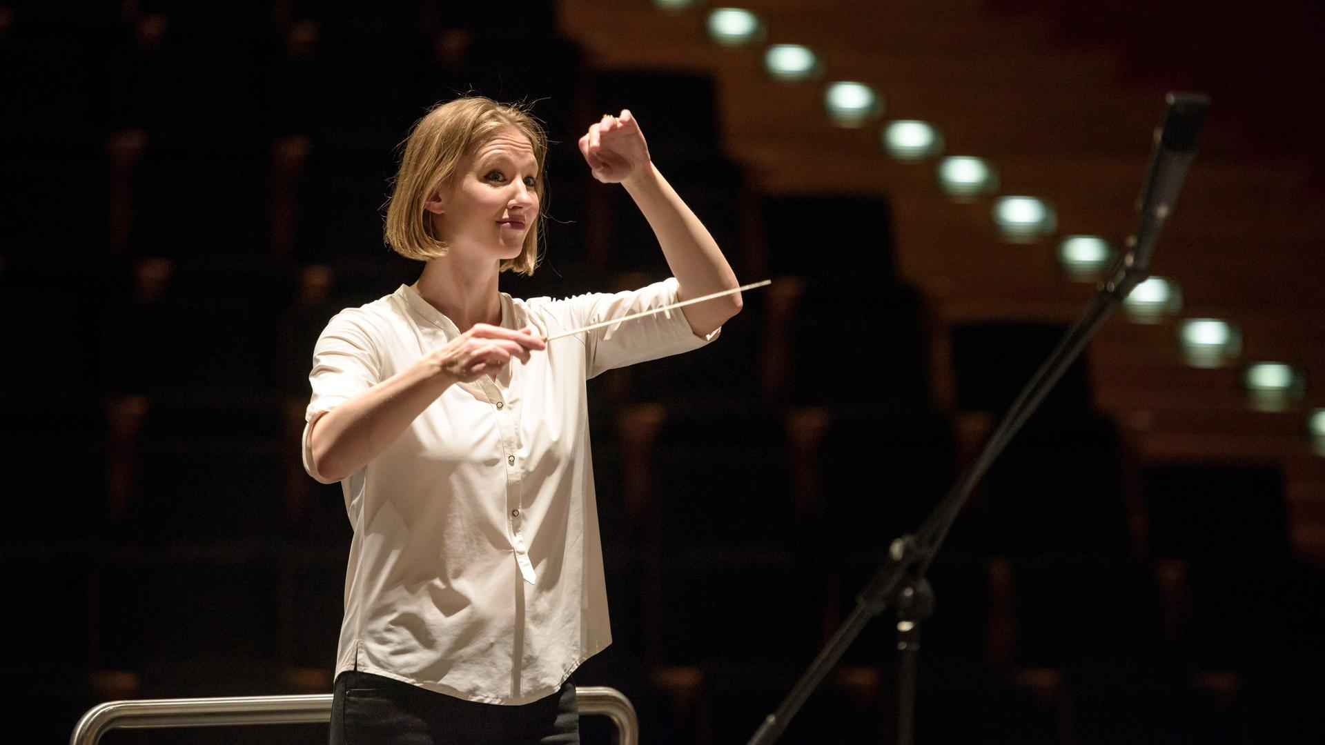 Eine junge Frau mit blonden kinnlangen Haaren blickt erwartungsvoll. Sie hat in der rechten Hand einen Taktstock, die linke hebt sie, um dem Orchester ein Zeichen zu geben. Es ist die Dirigentin Joana Mallwitz.