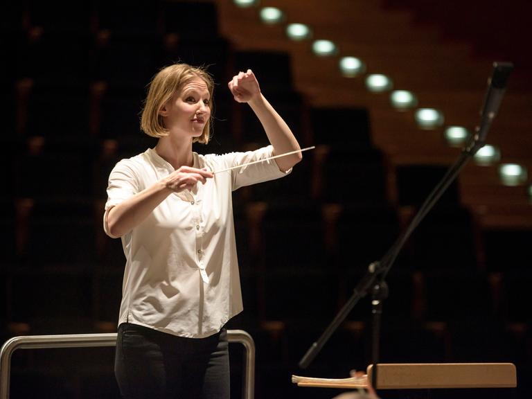 Eine junge Frau mit blonden kinnlangen Haaren blickt erwartungsvoll. Sie hat in der rechten Hand einen Taktstock, die linke hebt sie, um dem Orchester ein Zeichen zu geben. Es ist die Dirigentin Joana Mallwitz.