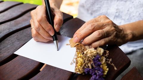 Die Mutter berichtete in Briefen an ihren erwachsenen Sohn vom Leben im Dorf. Zu sehen: Auf einem Tisch ein Blatt Papier, zwei Frauenhände, eine hält einen kleinen Strauß mit Lavendel, in der rechren Hand einen Kugelschreiber. 