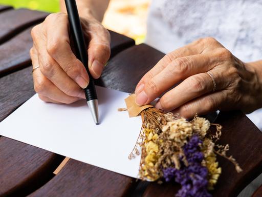Die Mutter berichtete in Briefen an ihren erwachsenen Sohn vom Leben im Dorf. Zu sehen: Auf einem Tisch ein Blatt Papier, zwei Frauenhände, eine hält einen kleinen Strauß mit Lavendel, in der rechren Hand einen Kugelschreiber. 