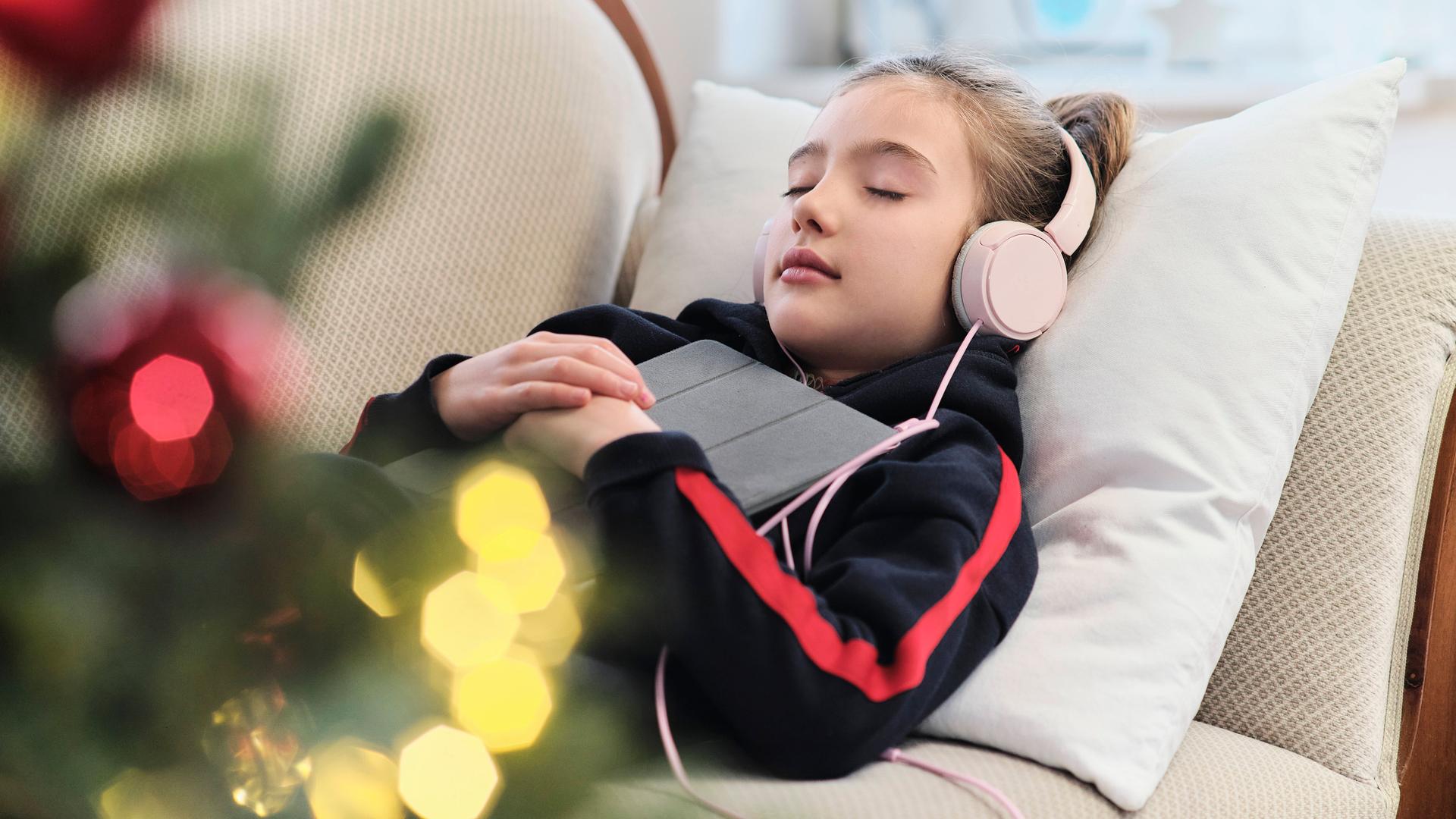 Ein Mädchen liegt entspannt mit Tablet und Kopfhörern auf dem Sofa. Im Vordergrund ist unscharf ein geschmückter Weihnachtsbaum zu sehen.