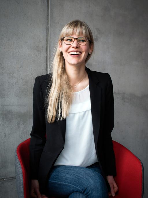 Die Psychologie-Professorin Eva Asselmann steht vor einer grauen Betonwand und guckt lachend zur Seite aus dem Bild hinaus.