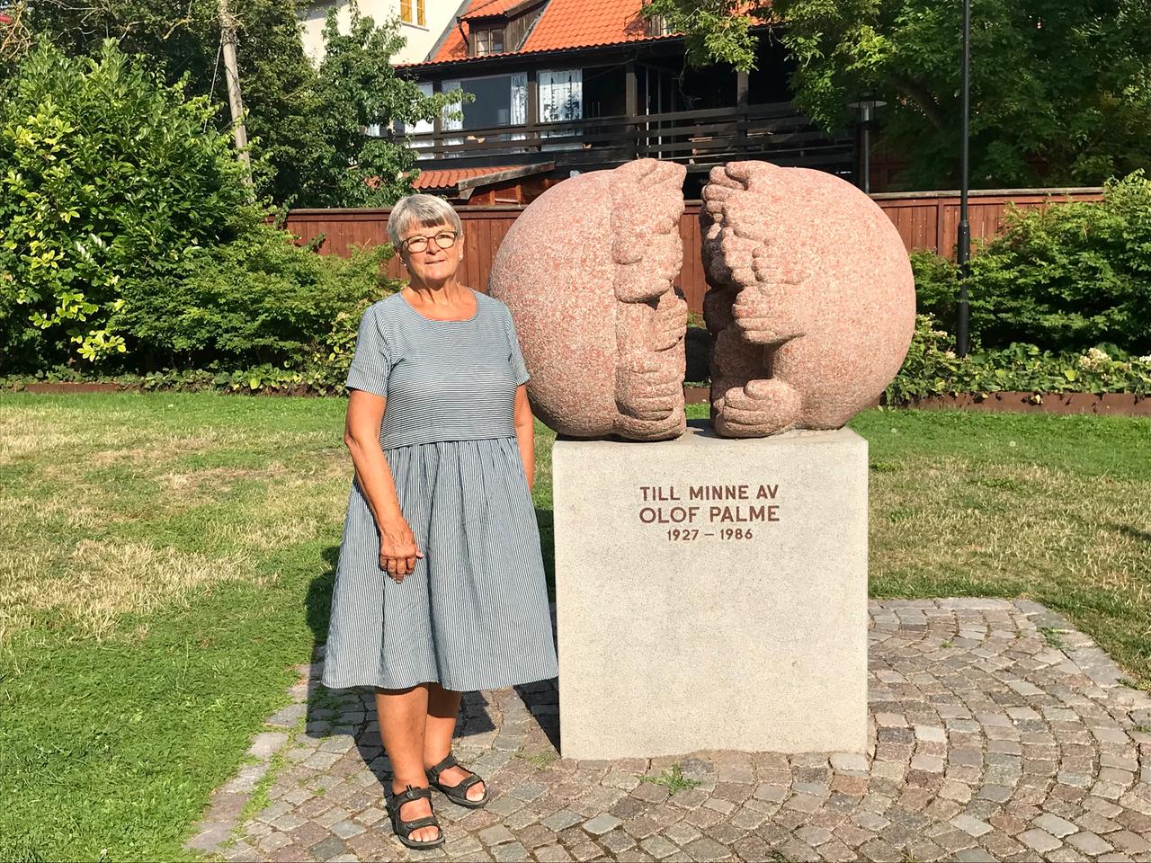 Eine ältere Dame im gestreiften Kleid steht auf einem kleinen Platz im Grünen neben einem Denkmal. Eine ältere Dame im gestreiften Kleid steht auf einem kleinen Platz im Grünen neben einem Denkmal.